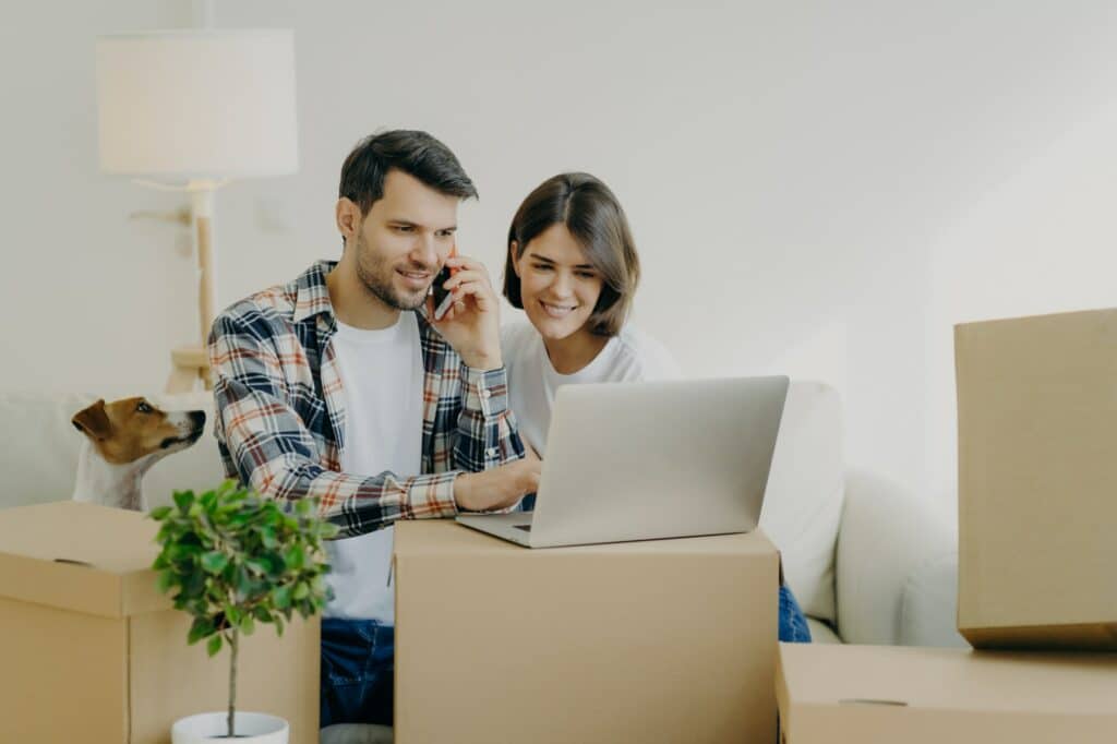 Happy man browses laptop in new flat, calls via smartphone, move in new apartment together with wife